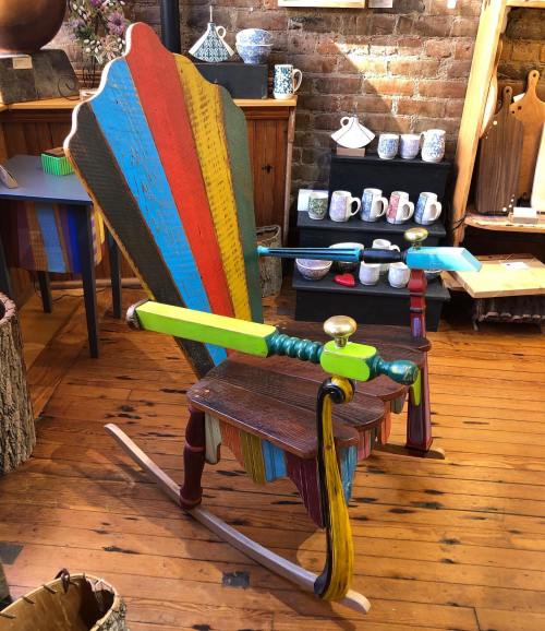 A colorful, handcrafted rocking chair with a striped backrest, set in a rustic shop with various pottery items.