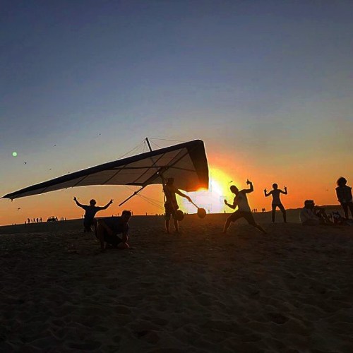 Silhouettes of people at the beach with a hang glider against a vibrant sunset sky.
