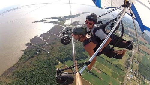 Two people hang gliding over a scenic landscape with water and fields below, enjoying an aerial adventure.