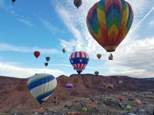 A colorful array of hot air balloons floats over a scenic landscape with hills and a blue sky.