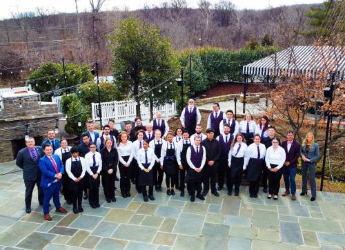 A large group of staff members in formal attire poses together outdoors at a venue with greenery and stone pathways.