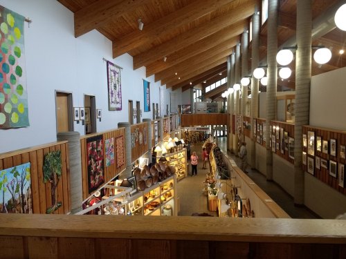 A spacious art gallery with wooden walls, colorful artwork, and visitors browsing displays under bright lights.