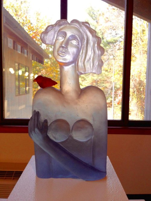 A translucent glass sculpture of a woman with a rose, set against a window with autumn foliage outside.