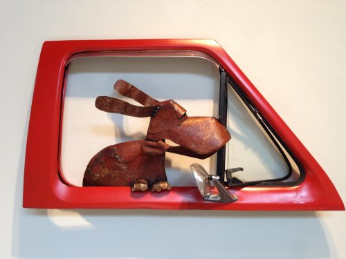 A whimsical sculpture of a dog made from metal, peeking out of a red car window frame.