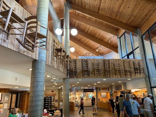 Interior of a spacious building with wooden beams, featuring a unique display of chairs on a balcony and people walking below.