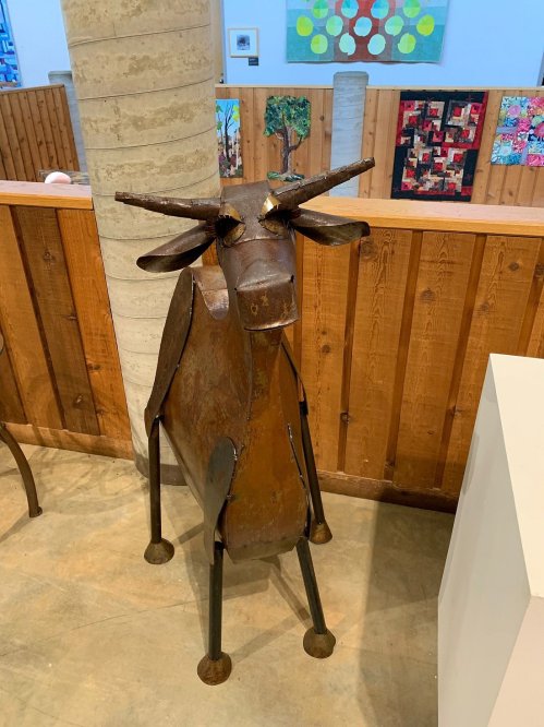 A metal sculpture of a cow stands in an art gallery, surrounded by wooden walls and colorful artwork in the background.