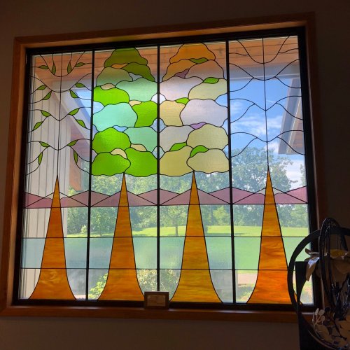 Stained glass window featuring colorful trees, clouds, and mountains, framed by a wooden border.