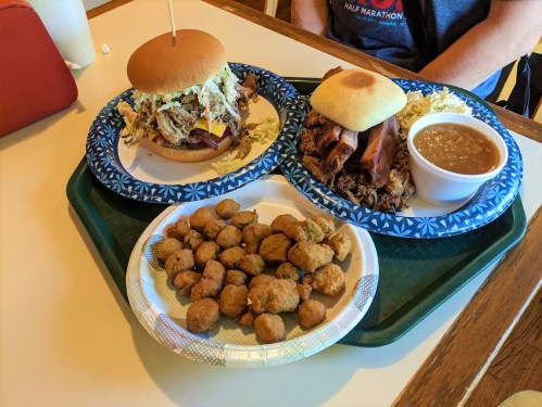A tray with two plates: one with a sandwich and pulled pork, the other with a bowl of beans and a side of fried okra.
