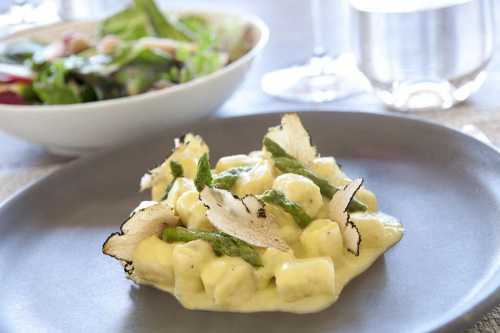 A plate of creamy gnocchi topped with asparagus and crispy vegetable slices, with a salad and water in the background.