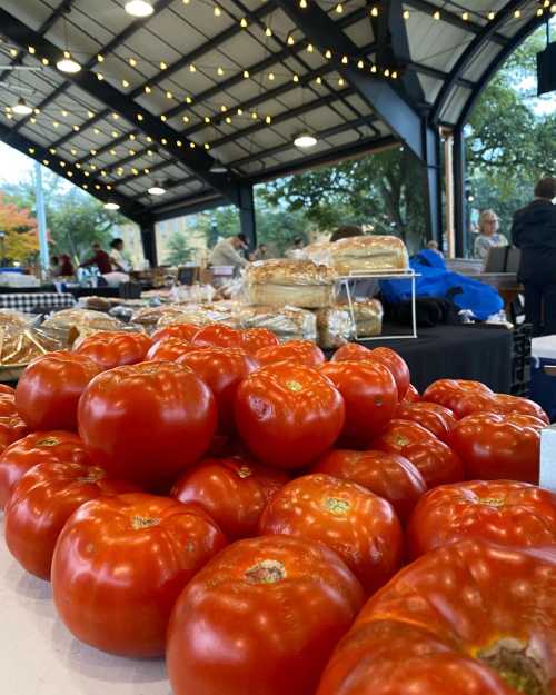 Fresh red tomatoes stacked in front of a bustling market scene with people and baked goods in the background.