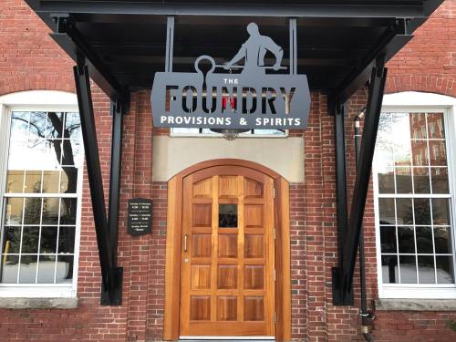 Sign for "The Foundry Provisions & Spirits" above a wooden door, set against a brick wall.