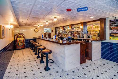 A cozy bar area with a jukebox, stools, and a variety of drinks displayed on shelves, featuring vintage decor.