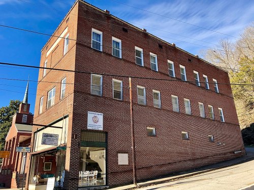 A brick building with multiple windows, located on a sloped street, with a church visible in the background.