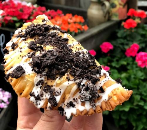 A hand holding a pastry topped with crushed cookies and icing, set against a backdrop of colorful flowers.