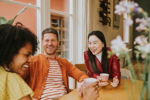 Three friends enjoying coffee together, laughing and chatting at a cozy table surrounded by plants and flowers.
