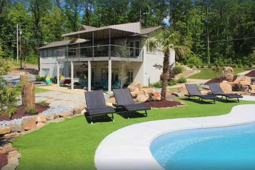 A modern house with a pool, surrounded by rocks and greenery, featuring lounge chairs on a grassy area.