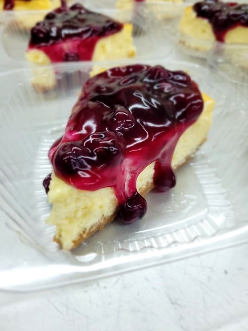 A slice of cheesecake topped with a glossy berry sauce, served in a clear plastic container.