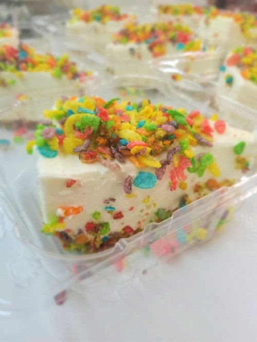 A close-up of a dessert slice topped with colorful cereal sprinkles in a clear plastic container.