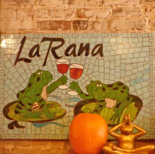 A colorful mosaic sign reading "La Rana" featuring two frogs toasting with glasses, alongside a pumpkin and a statue.