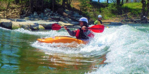 A person kayaking on a river, creating splashes, with another kayaker in the background among trees and rocks.