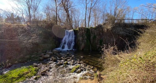A serene waterfall cascades over rocks, surrounded by trees and greenery under a clear blue sky.