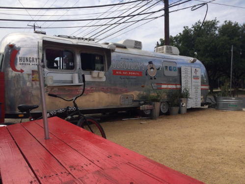 A silver food truck with a sign reading "Sourdoughs" parked in a gravel area, surrounded by trees and power lines.