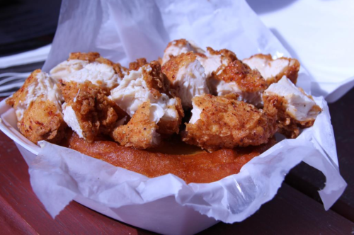 A serving of crispy fried chicken pieces on a bed of orange cornbread, served in a white paper container.