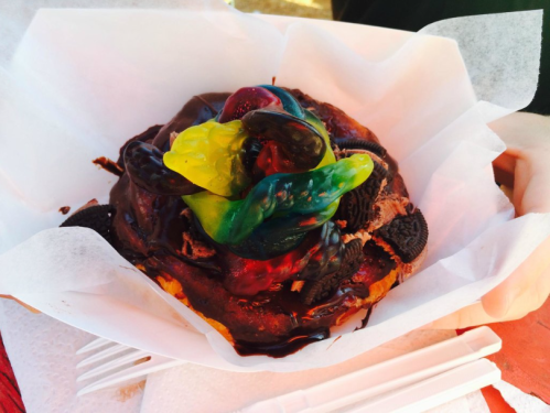 A chocolate-covered pastry topped with gummy candies and crushed cookies, served on a white paper wrapper.