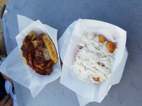 A plate with a bacon sandwich and a dessert topped with icing and small dough balls, both served in paper trays.