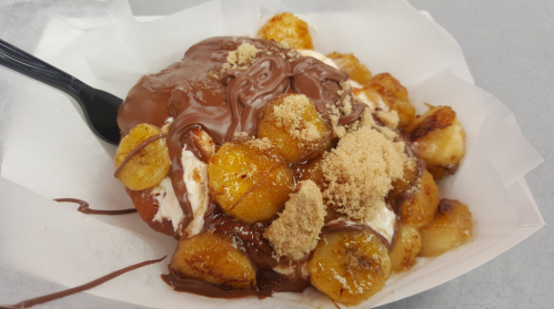 A dessert bowl featuring fried bananas topped with chocolate sauce, cream, and brown sugar, served in a paper container.