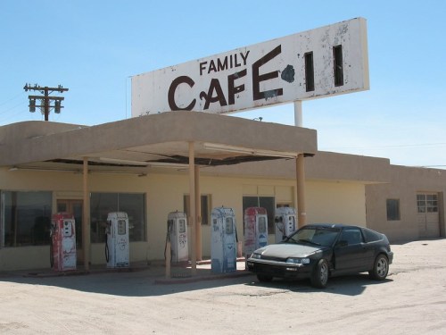 Abandoned gas station and café with vintage pumps and a parked car under a clear blue sky.
