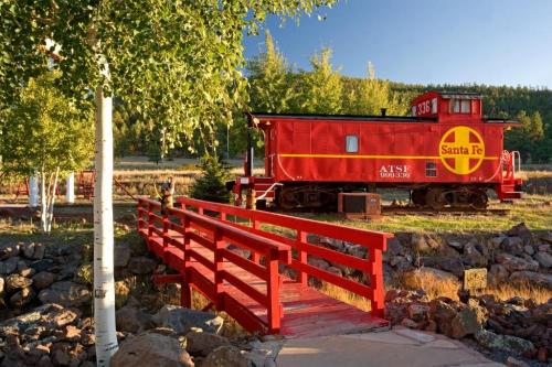 A red Santa Fe caboose beside a wooden bridge, surrounded by trees and rocky terrain in a scenic landscape.