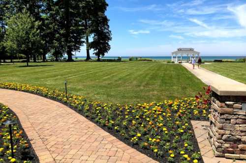 A scenic view of a manicured lawn with a flower-lined path leading to a gazebo by the water under a clear blue sky.