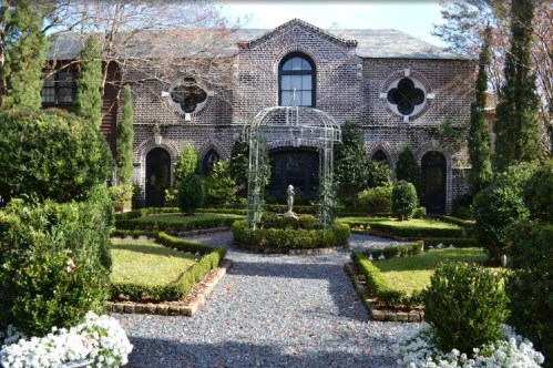 A charming brick house surrounded by manicured gardens, featuring a decorative gazebo and lush greenery.