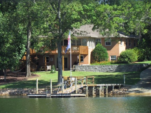 A brick house with a deck overlooks a calm lake, featuring a small dock and lush greenery surrounding the property.