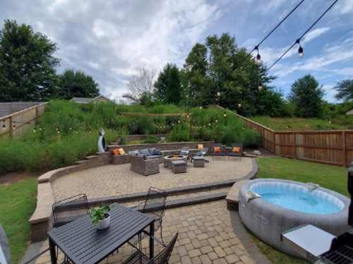A cozy backyard with a stone patio, seating area, fire pit, hot tub, and greenery under a cloudy sky.