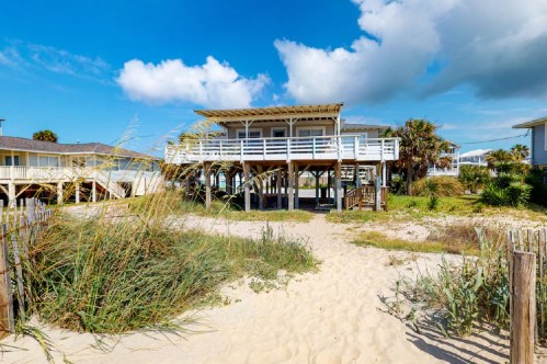 A beach house on stilts surrounded by sandy dunes and tall grass under a blue sky with fluffy clouds.