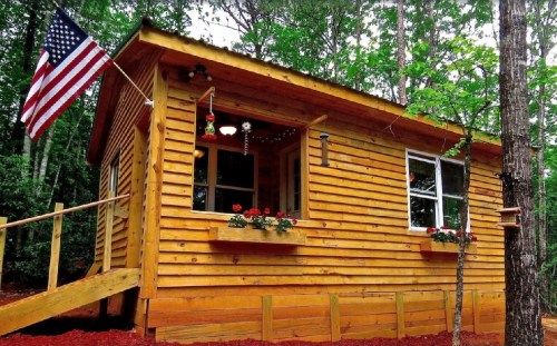 A cozy wooden cabin surrounded by trees, featuring an American flag and flower boxes under the windows.