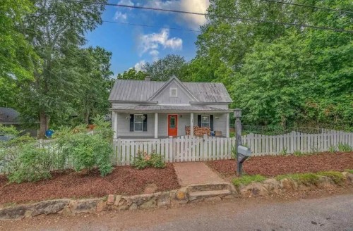 A charming gray house with a metal roof, orange door, and white picket fence, surrounded by greenery and a gravel path.