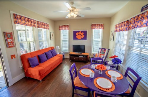 Bright living room with orange couch, purple dining set, and Clemson-themed decor, featuring large windows and natural light.