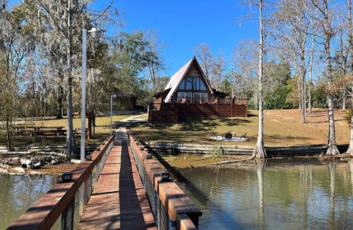 A wooden bridge leads to a modern A-frame cabin by a calm lake, surrounded by trees and clear blue skies.