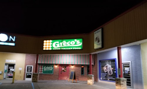 Exterior of Greco's restaurant at night, featuring a bright sign that reads "Good Italian Food" and striped awning.