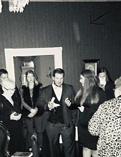 A group of people in formal attire gathered in a dimly lit room, with one person gesturing while others watch.