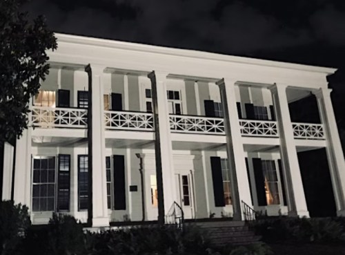 A historic white house with columns, dark windows, and a staircase, illuminated at night against a cloudy sky.