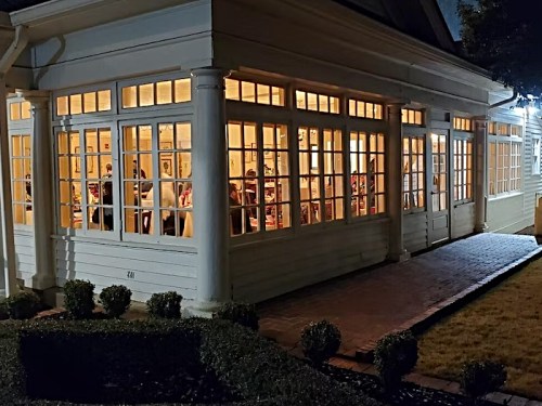 A well-lit building with large windows, showing people inside, surrounded by neatly trimmed hedges at night.