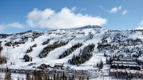 Snow-covered mountains with ski slopes, a lodge, and a parking area under a blue sky with scattered clouds.
