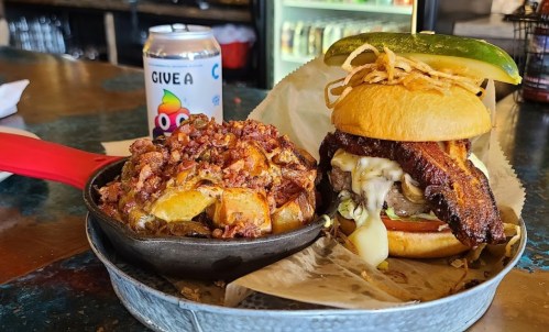 A hearty burger topped with pickles and crispy onions, served with a side of loaded potatoes and a can of soda.