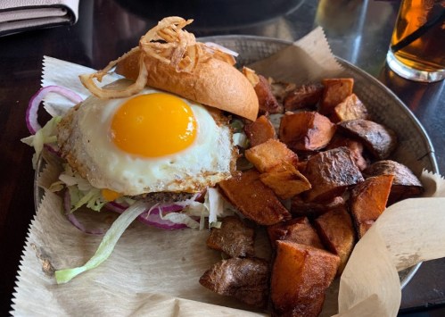 A plate featuring a sandwich with a fried egg, lettuce, and crispy onions, served with seasoned potato wedges.