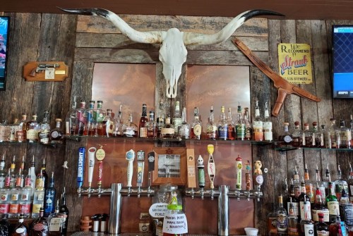 A rustic bar display featuring a longhorn skull, various liquor bottles, and beer taps against a wooden backdrop.