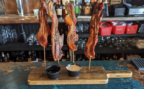 Three skewers of crispy bacon on a wooden board, served with two dipping sauces in small cups.